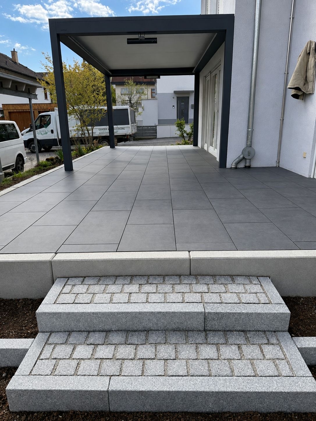 Terrasse mit grauen Feinsteinzeugplatten, Granitstufen und Pergola am Haus